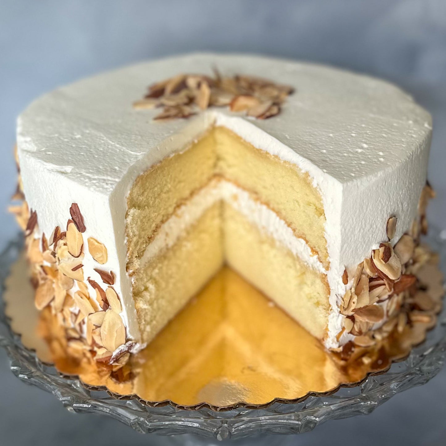 A slice of almond cream cake with white frosting and almonds on top, placed on a clear plate.